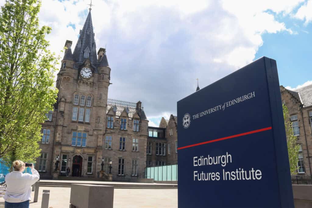 A modern blue sign with white and red text reads "Edinburgh Futures Institute, The University of Edinburgh." A historic building with a clock tower stands in the background under a cloudy sky. A person is taking a photo on the left near green trees.