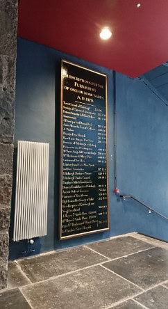 A tall signboard with gold text in a black frame is mounted on a blue wall next to a white radiator. The board lists various rules or information. The floor consists of gray stone tiles and a metal handrail is attached to the wall on the right.