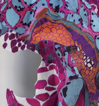 A close-up of an intricately patterned fabric with vibrant colors including purple, blue, and orange. The design features abstract shapes and textures with a partially torn section on the left side.