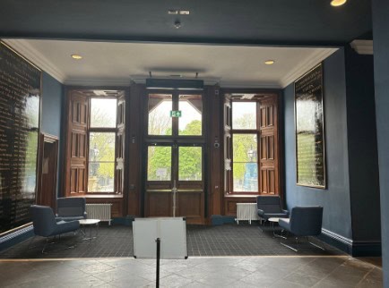 A spacious indoor lounge area with large wooden-framed windows allowing natural light to pour in. There are four blue cushioned chairs around the room, and two large, black etched panels on either side of the entrance. A blank white sign is in the center foreground.