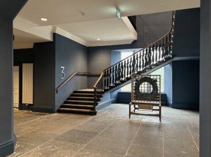 A modern interior with dark walls and stone flooring features a wide staircase with wooden railings leading to an upper level. There is a large "3" on the staircase wall, and a decorative metal frame displayed on the right. Natural light comes through a window.