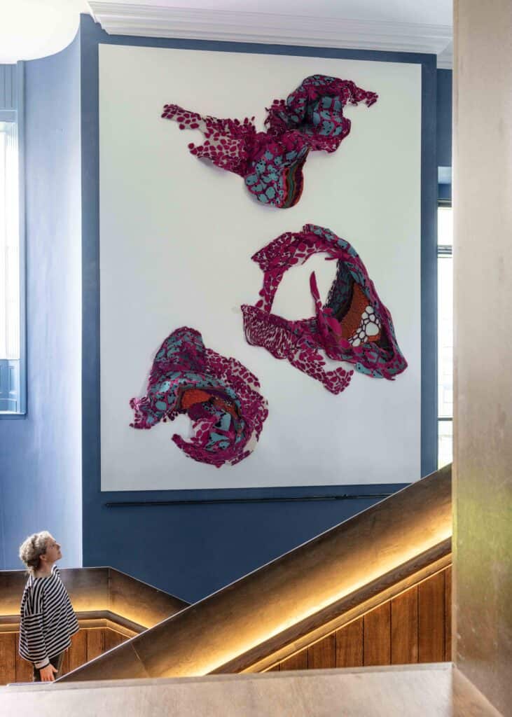 A person with curly hair wearing a black-and-white striped shirt gazes up at an abstract wall art installation featuring three vibrant, multi-colored textile pieces hung on a white background. The scene is located beside a wooden stairway with a blue wall.