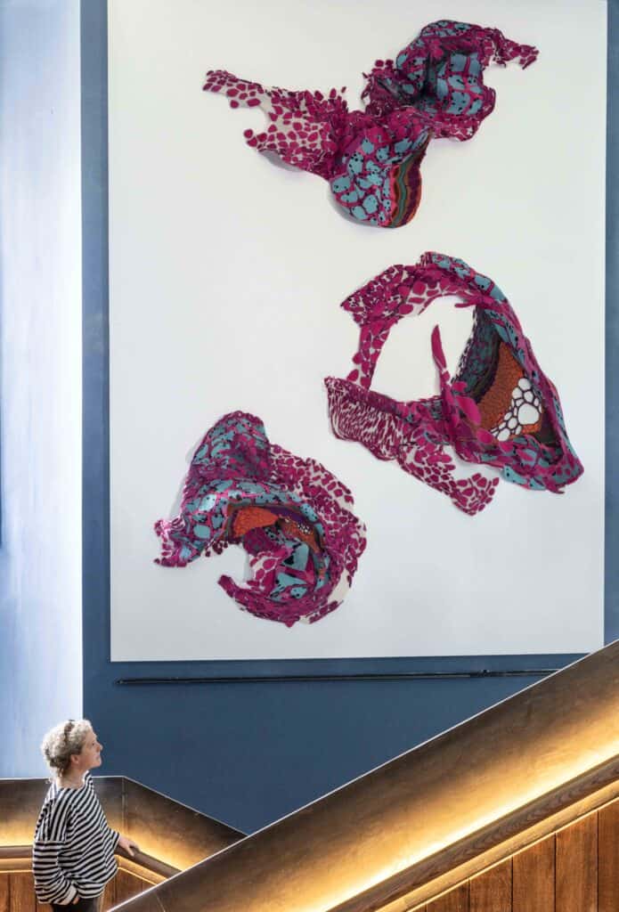 A person with blonde hair in a patterned blouse looks up at a large, vibrant textile art piece on a wall in a modern interior. The artwork consists of abstract, colorful fabric shapes in shades of purple, pink, blue, and red. They are mounted on a white canvas.