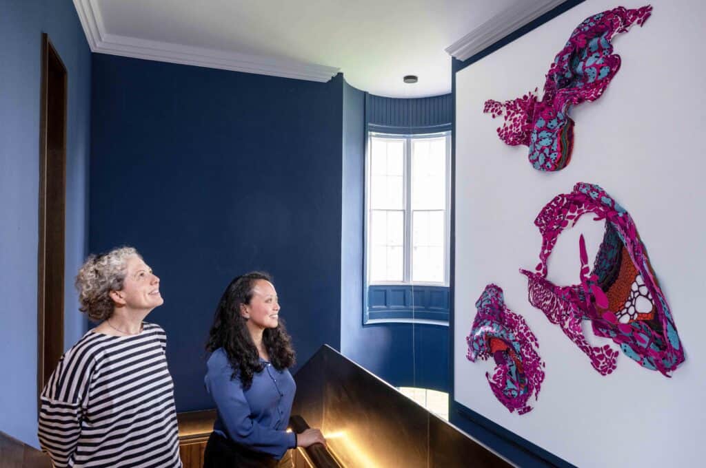 Two individuals are looking at a large, colorful, abstract art piece mounted on the wall in a modern, blue-painted room. The artwork features vibrant pink, red, and blue patterns. Light streams in from a window, illuminating the space and the artwork.