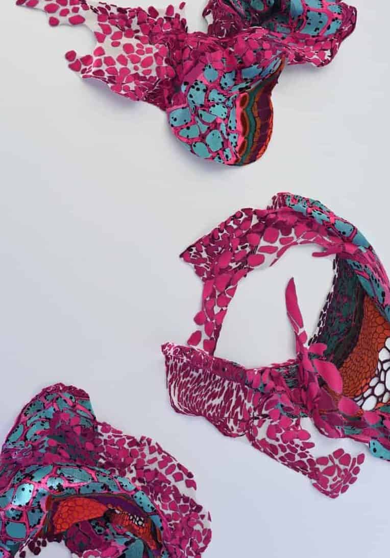 Brightly colored fabric scarves with vibrant patterns, including pink and turquoise hues, are arranged randomly on a white background. Some are loosely tied, showcasing various textures and designs.