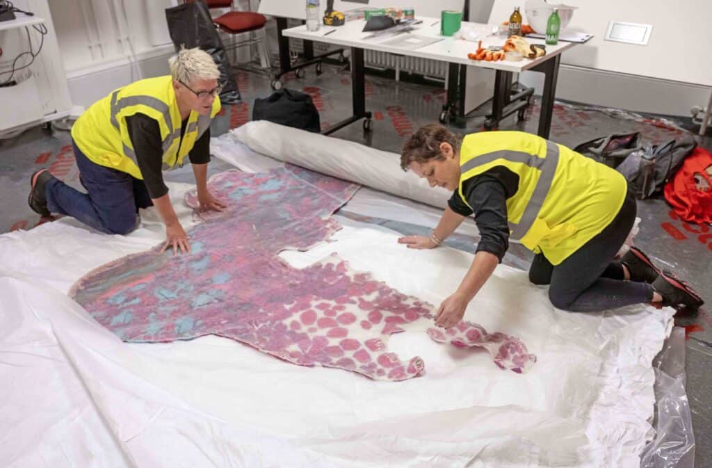 Two people, wearing yellow safety vests, are carefully handling a large, abstract, multi-colored fabric piece spread out on the floor. They appear to be in a workspace with tables and chairs covered with various tools and materials in the background.