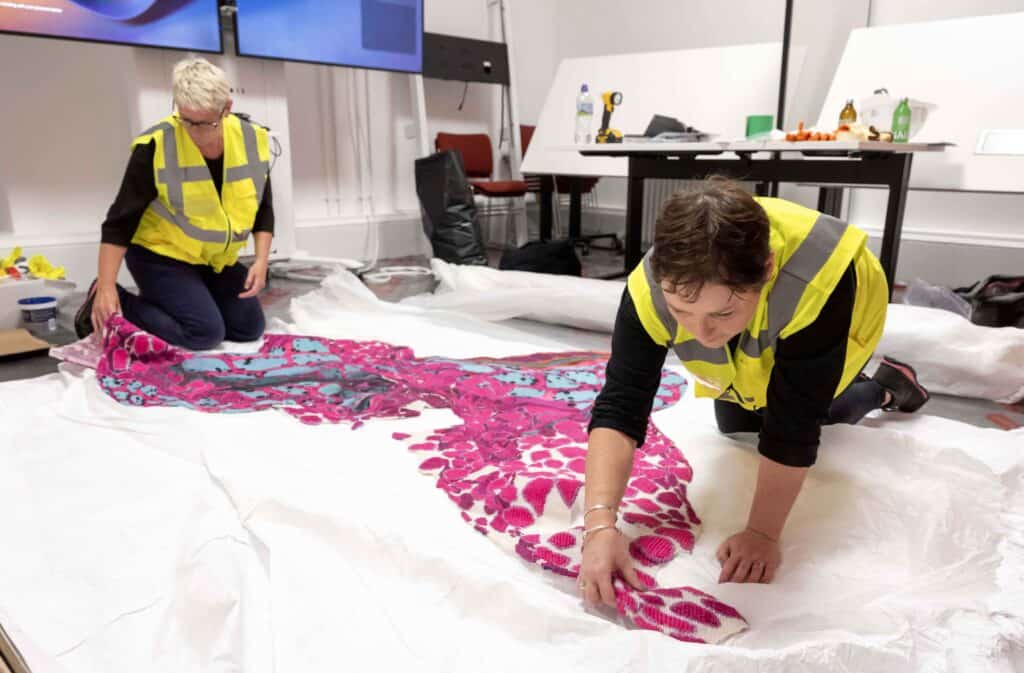 Two people wearing high-visibility vests are carefully arranging a large, colorful textile artwork on the floor. The textile features vibrant pink and blue patterns. Various tools and supplies are scattered on a nearby table and chair.