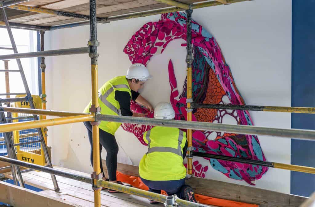 Two construction workers in yellow safety vests and white helmets are installing a vibrant, abstract mural with shades of pink, red, and blue on a white wall. They are surrounded by scaffolding, indicating they are working in a partially unfinished space.