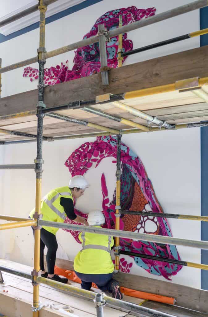 Two construction workers in high-visibility vests and white hard hats work on installing a colorful, abstract wall art piece while standing on scaffolding. The artwork features a large, intricate design in red, pink, and blue hues.