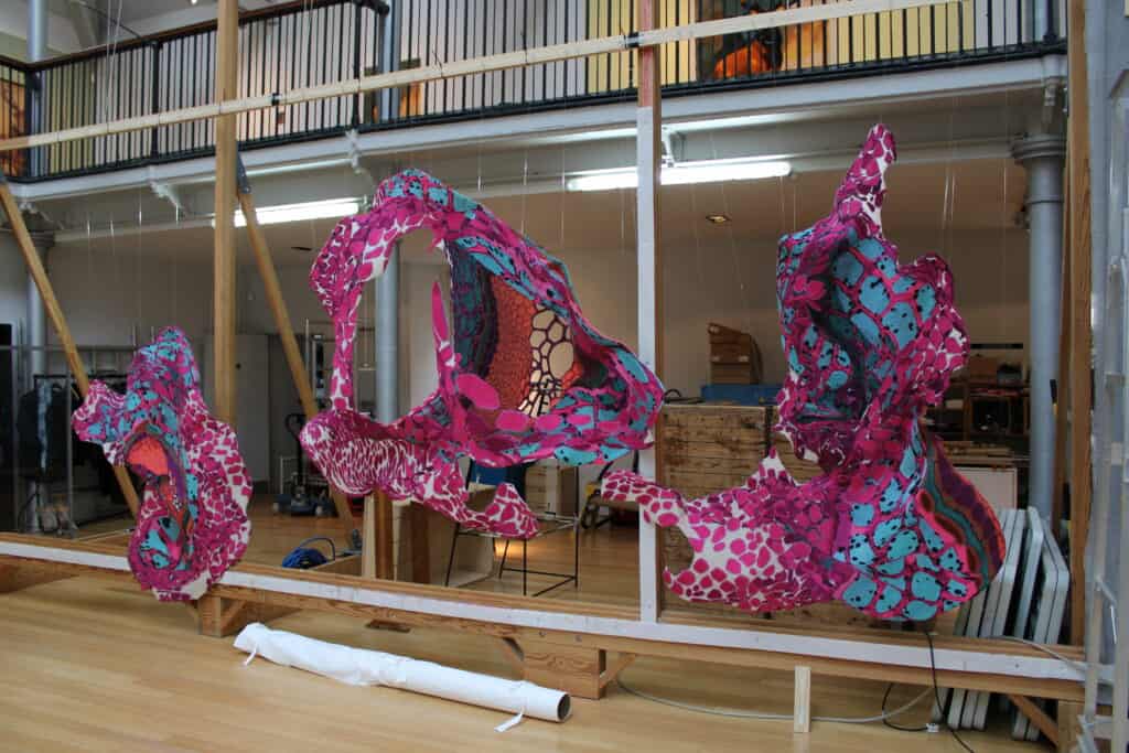 An art installation featuring vibrant, abstract fabric sculptures in shades of pink, blue, and red. The fabric pieces are suspended from the ceiling in an indoor space with a wooden floor, metal railing, and various studio equipment in the background.