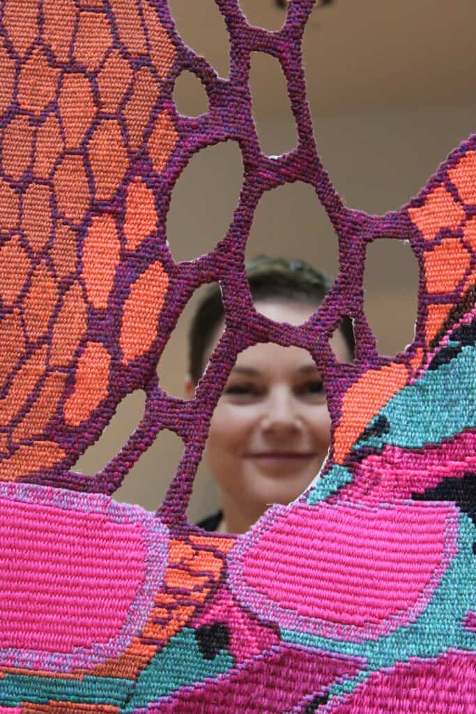 A person with a friendly expression looks through an intricate and brightly colored textile art piece featuring patterns in pink, purple, and teal. The textile has an organic, web-like design with small and large holes.