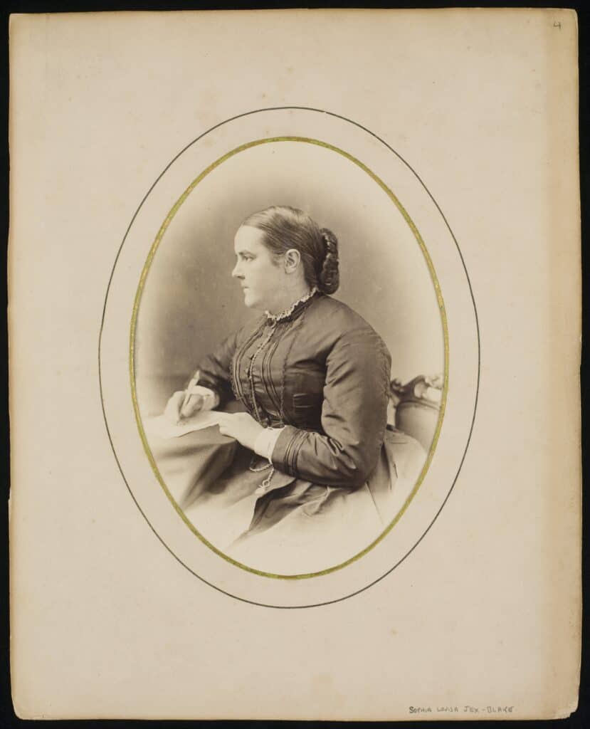 A sepia-toned vintage photograph of a woman in profile, seated and holding a piece of paper. She wears a long-sleeved dress with lace details. The photo is framed in an oval shape on a plain background with handwritten text at the bottom right corner.