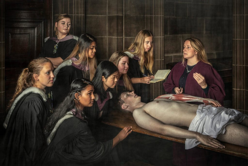 A group of female students in graduation gowns observe a teacher conducting a lesson with a lifelike model of a body on a table. The setting appears to be an old, wood-paneled room. One student is taking notes. The scene resembles a historical anatomy class.