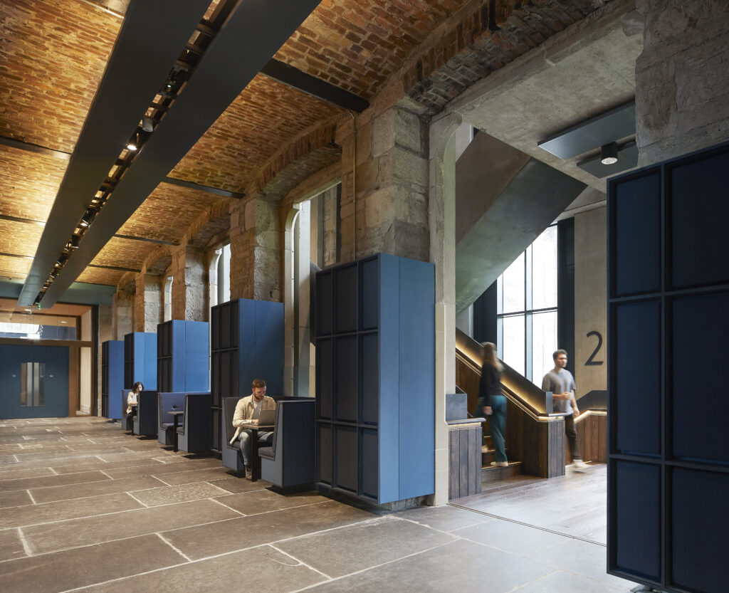 A modern interior features tall brick walls and large windows. People are seated in private booth-like spaces along the left, with a staircase to the right. Blue accents and overhead lighting create a contemporary atmosphere.