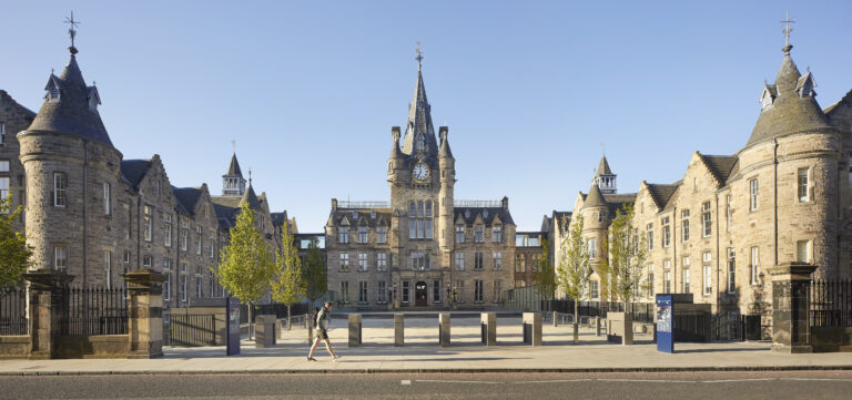 A grand, historic stone building with a central clock tower and symmetrical turrets on each side. A person walks on the sidewalk in front, and there are small trees lining the walkway. The sky is clear and blue.