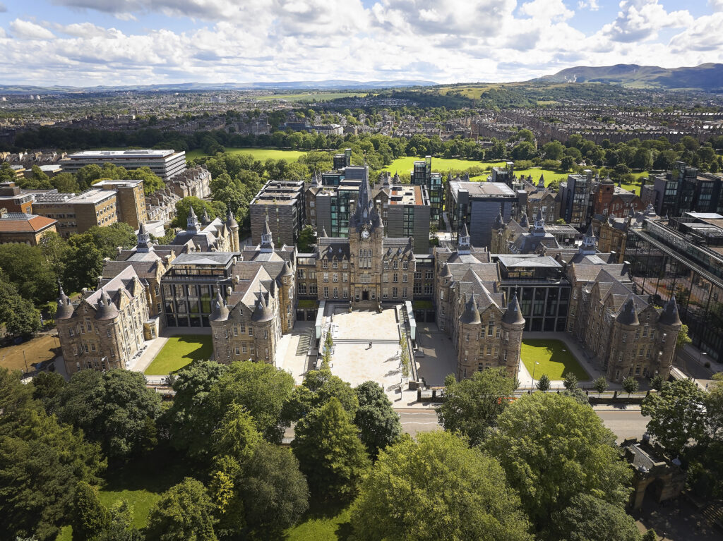 Aerial view of a historic, grand building with spires and a courtyard, surrounded by lush green trees. In the background, a cityscape extends toward distant hills under a partly cloudy sky.