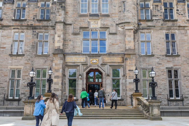 People walking towards an ornate stone building with large windows and two lamp posts flanking the entrance. The building has inscriptions of years and Latin text above the door, indicating historical significance.