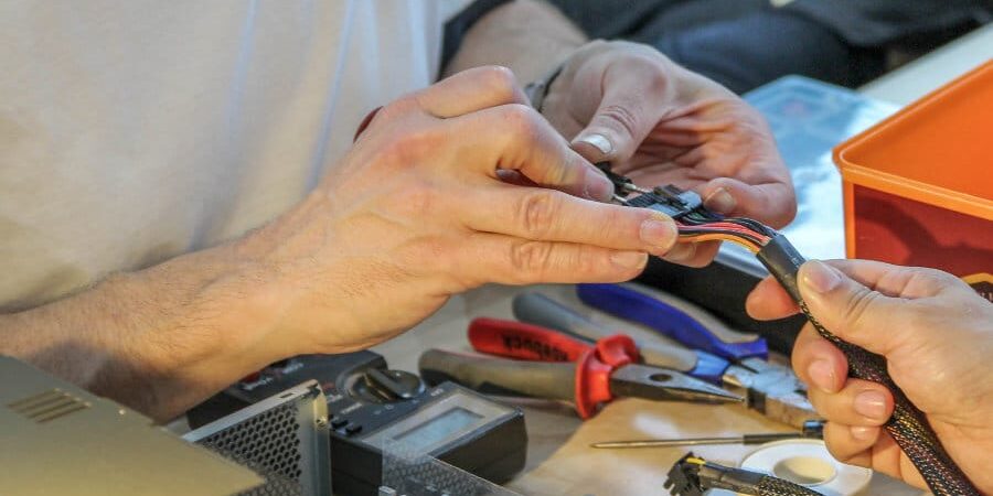 Two people work together on assembling or repairing electronic equipment. They handle colorful wires surrounded by tools, including a screwdriver and pliers, at a cluttered workspace.