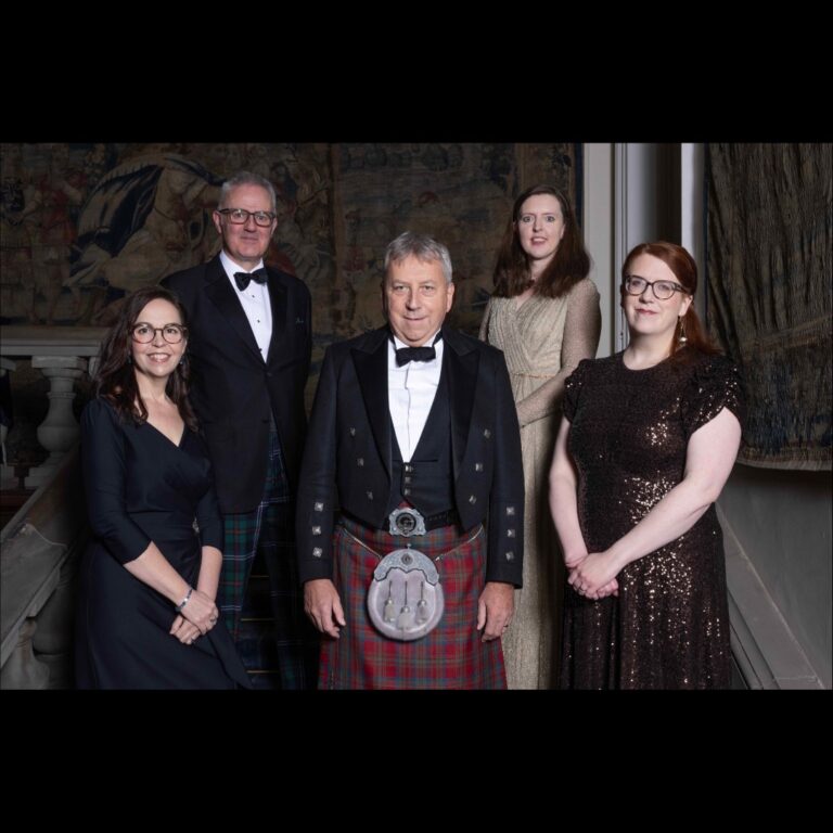 A group of five people dressed in formal attire, including kilts and dresses, standing on a staircase with a tapestry in the background. The setting appears elegant, suggesting a special occasion or event.