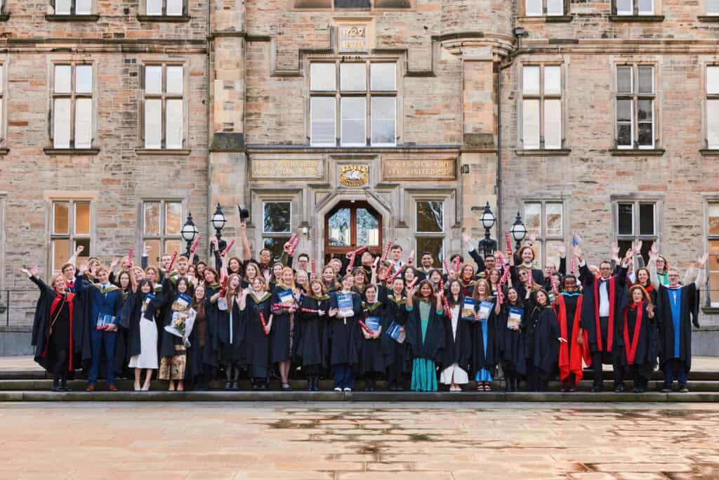 A large group of graduates in black gowns and caps celebrate in front of a historic building. Some are holding diplomas, and others are raising their arms in joy. The building facade features brickwork and large windows.