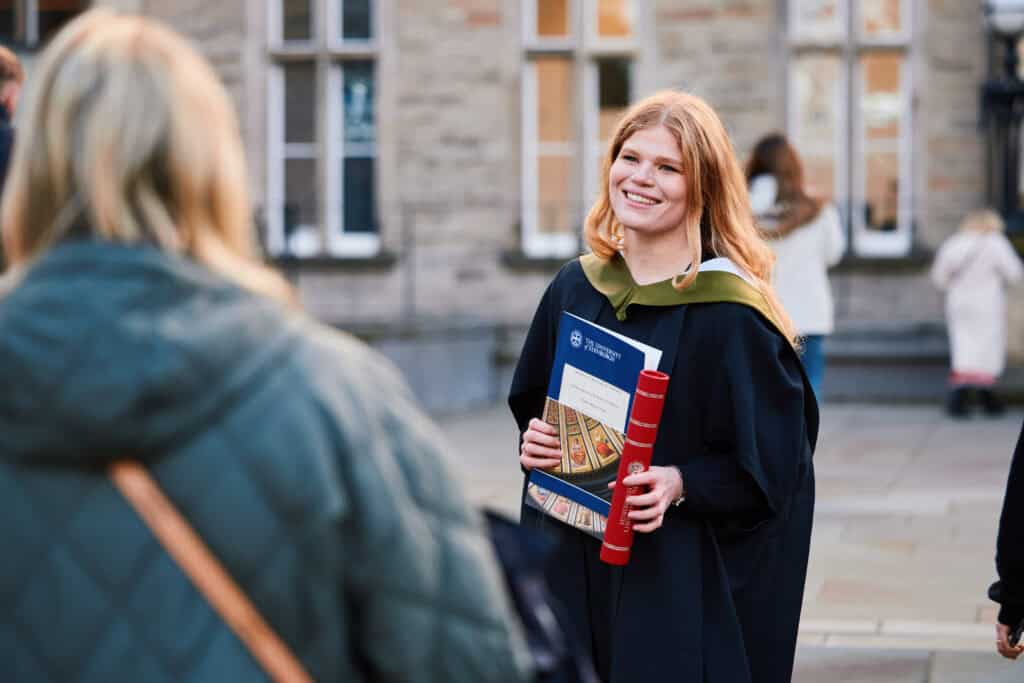 A graduate in a black gown and green hood smiles while holding a diploma and a certificate outdoors. Another person, slightly out of focus, stands nearby. The background features a brick building with large windows.