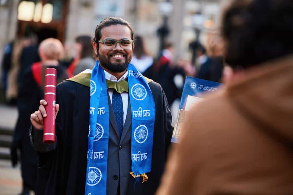 A graduate in a cap and gown holds a diploma and wears a blue stole with white patterns. He smiles outdoors, where people are gathered in the background, suggesting a graduation ceremony.