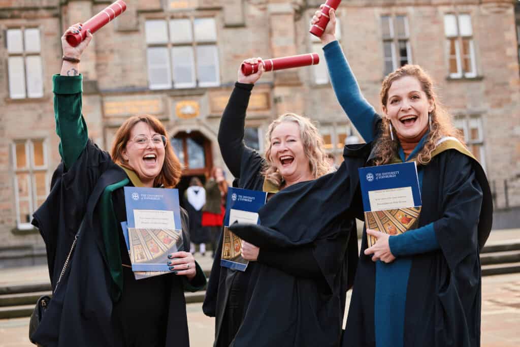 Three smiling graduates wearing gowns and holding diplomas in the air celebrate in front of a historic building. They hold brochures from the University of St Andrews.