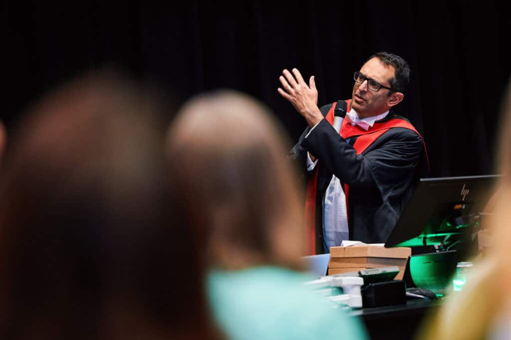 A man in a graduation gown and glasses speaks passionately in front of a lectern. He gestures with one hand, emphasizing his point. Blurred audience members sit in the foreground, and a dark backdrop adds contrast.