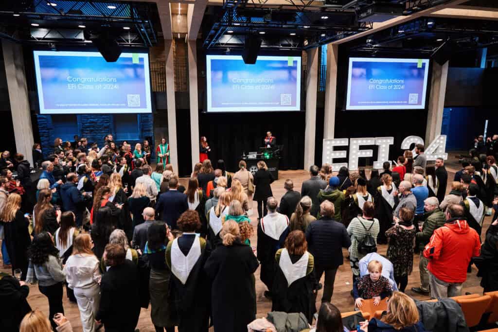 A large group of people, some wearing graduation attire, gather in a hall. Two screens display "Congratulations EFI Class of 2024." The setting is festive with a stage and a speaker addressing the audience.