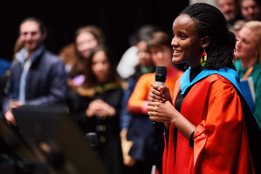 A person in a red academic gown and colorful earrings speaks into a microphone. They are smiling and standing on a stage, with a blurred audience in the background.