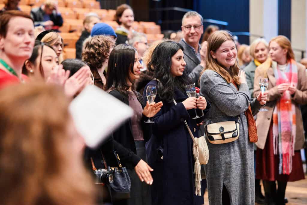 A group of people are standing and clapping in an auditorium. One person holds a glass of wine. They appear to be engaged and smiling, suggesting a celebratory or formal event. Seats in the background are partially filled.