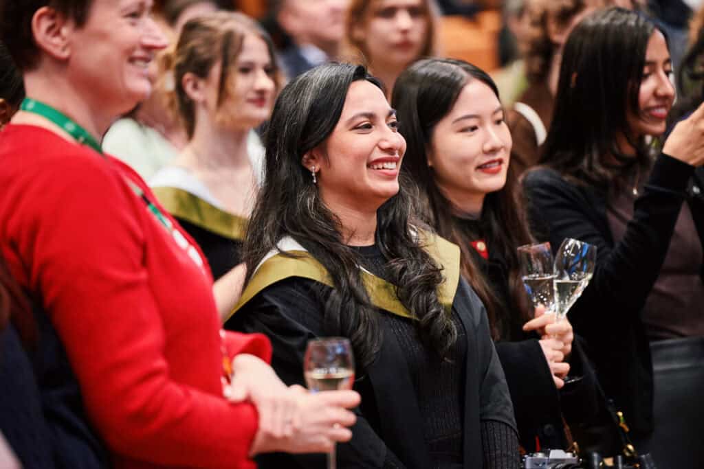 A group of people, mostly women, smiling and celebrating at a formal event. Some are wearing black outfits with green and gold scarves or stoles, and a woman in green holds a champagne glass. Their expressions are joyful and relaxed.