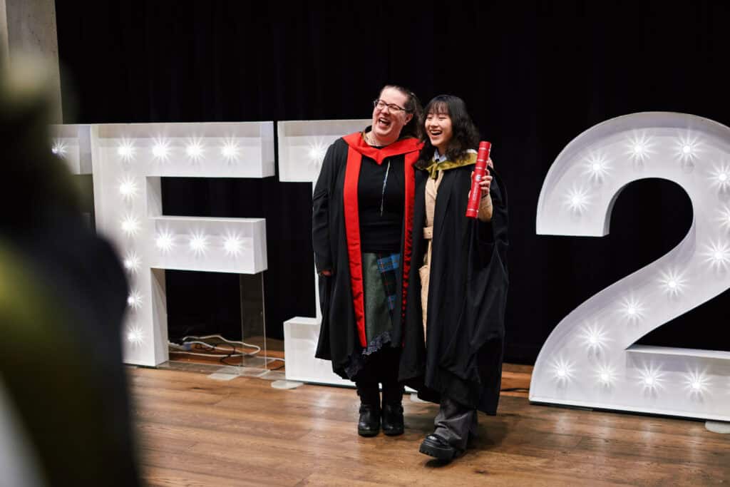 Two people in graduation attire stand smiling together in front of large illuminated letters and numbers. One holds a red diploma tube. Both appear joyful, celebrating an academic achievement.