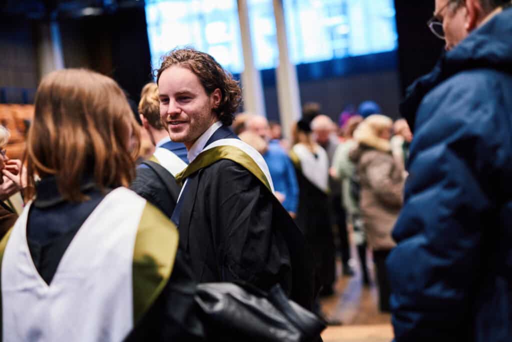 A group of people in graduation attire is gathered in a large hall. One person in the foreground, wearing a gown with a yellow and black hood, is turned and smiling. The background shows others in conversation, with some wearing winter coats.