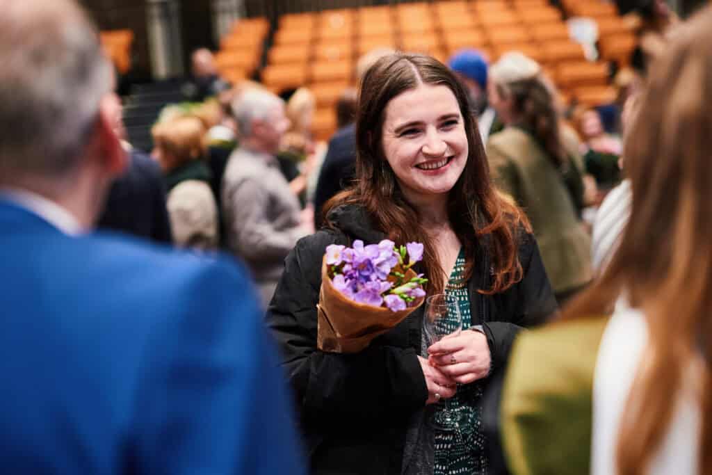 A woman smiling and holding a bouquet of purple flowers stands among a group of people in a large room with orange seating. She appears to be engaged in conversation.