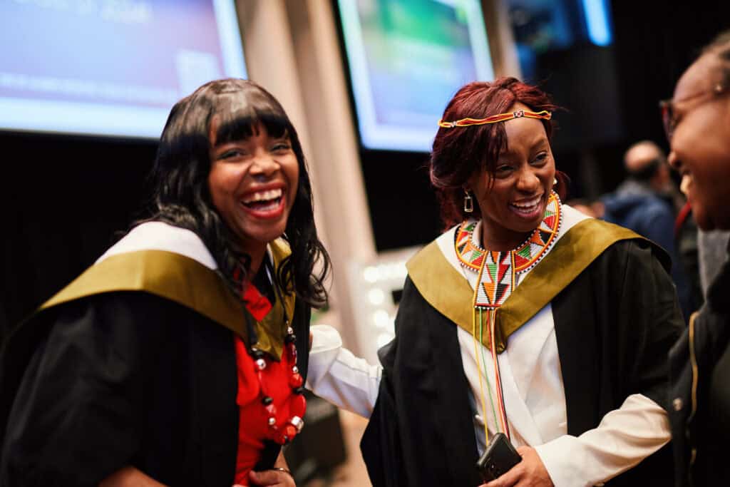 Two graduates celebrate at a ceremony, wearing academic gowns with gold accents. One wears a red dress and the other a white outfit with colorful beaded jewelry. Both are smiling and appear to be sharing a joyful moment.