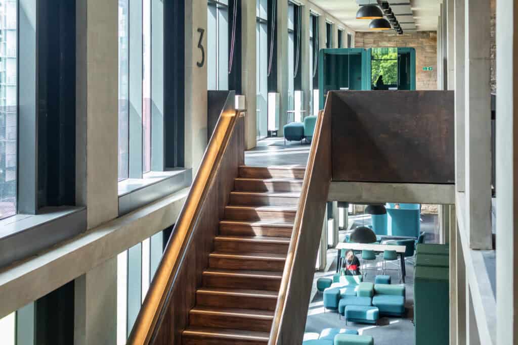 A modern interior with large windows, a wooden staircase leading to the upper level, and seating areas with turquoise chairs and tables. The sunlight brightens the space, creating a welcoming atmosphere.