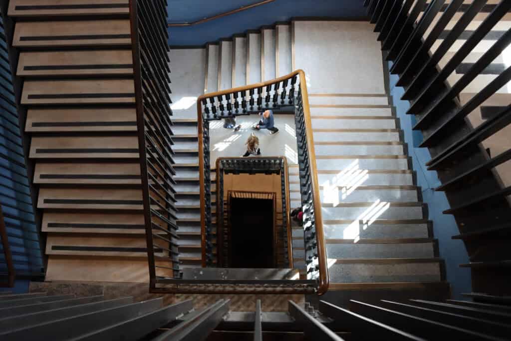 Aerial view of a multi-level staircase with a person sitting in the center on the landing. Sunlight casts shadows on the steps. The stair railings create geometric patterns, enhancing the perspective.