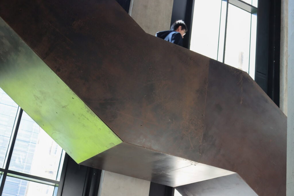 A person walking up a large, industrial-style metal staircase inside a modern building with tall windows. The staircase has a rusty brown texture and strong geometric lines. Bright natural light filters through the windows.