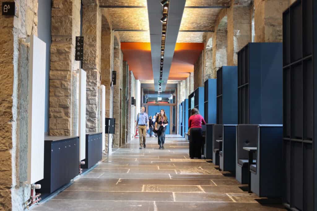 A modern hallway with stone walls and blue cubicles on the sides. Three people walk down the corridor, with two facing the camera. The ceiling features a center light strip, and the floor is made of large stone tiles.