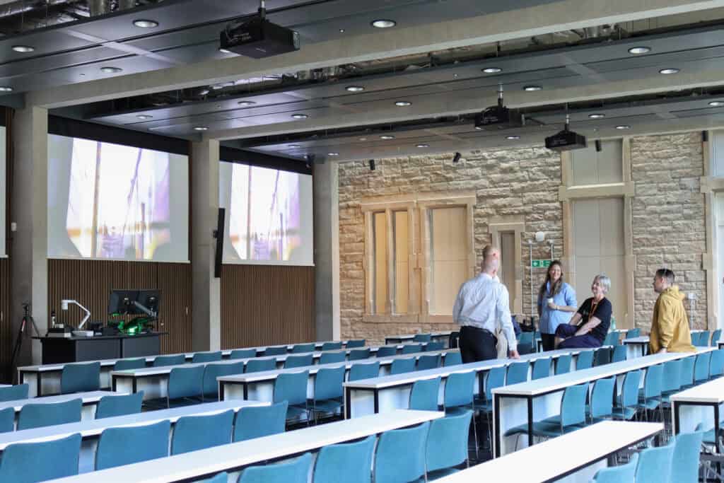 A modern lecture hall with empty blue and white seats arranged in rows. Four people are conversing at the front near a podium with computer equipment. Two large screens display images on the walls, and the room has a mix of wood and stone accents.