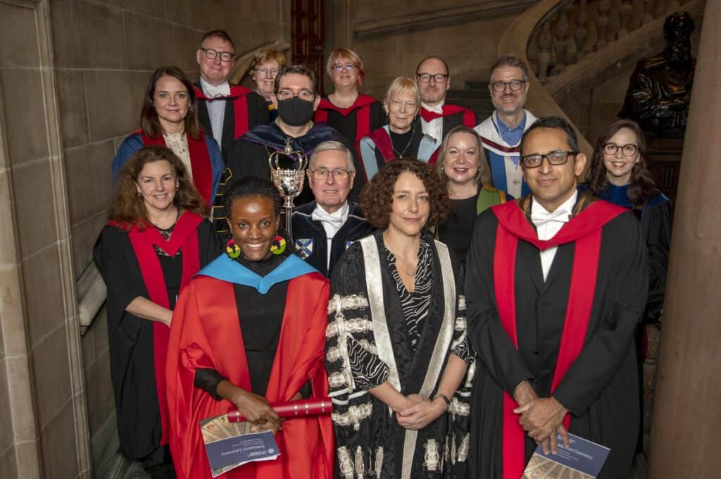 A group of people wearing academic regalia, including gowns and hoods, gathered indoors on a staircase. Some hold certificates or awards. They are smiling and appear to be at a formal academic event or ceremony.