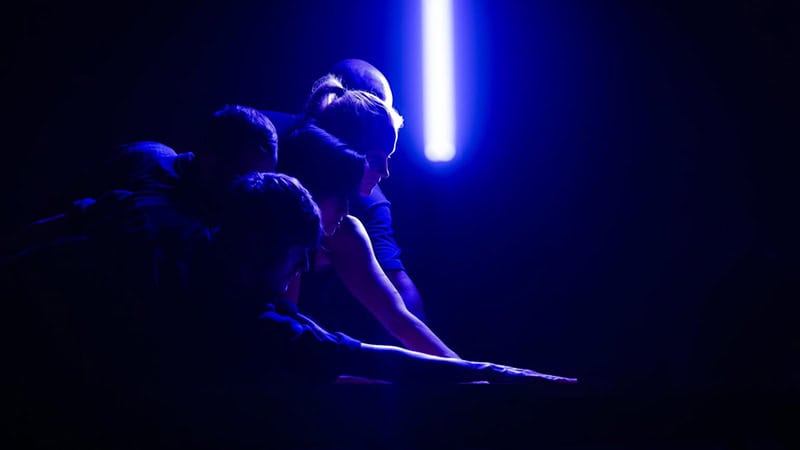 Four people lean forward in a dark space illuminated by a blue light. The light creates silhouettes and highlights their profiles, casting dramatic shadows.