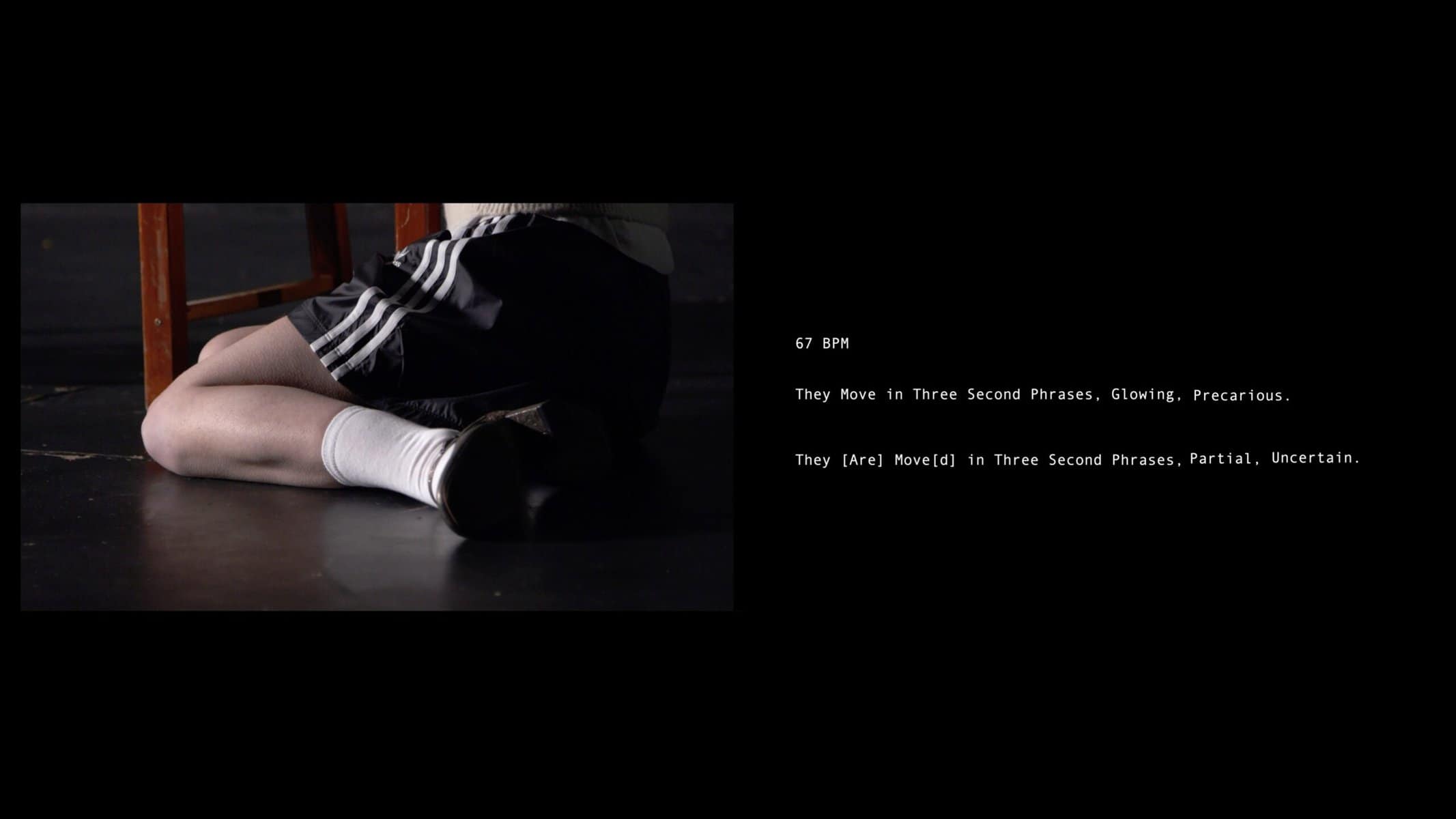 Georgia Gardner, Still from Body Memo (2023). The image shows a person in black shorts and white socks lying on the floor next to a stool. Text to the right reads: "67 BPM. They Move in Three Second Phrases. Glowing, Precarious. They [Are] Move(d) in Three Second Phrases. Partial, Uncertain.