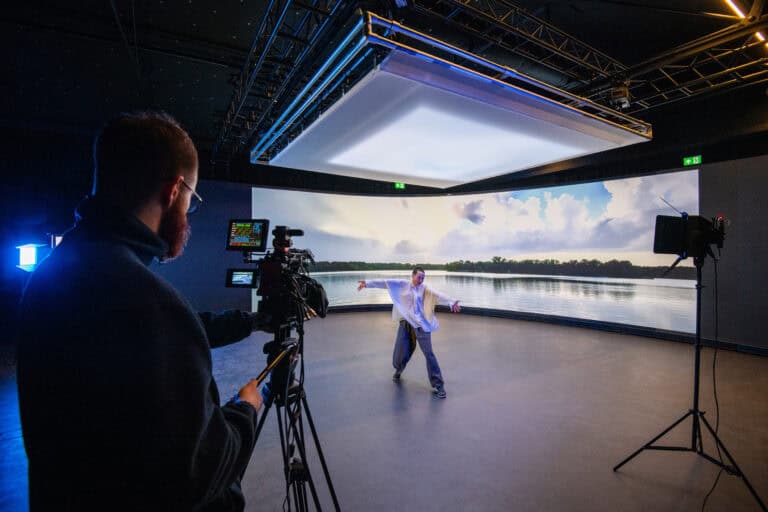 A person dances energetically in front of a large digital screen displaying a serene lake scene with clouds. A camera operator films the performance. Studio lights and equipment are visible.