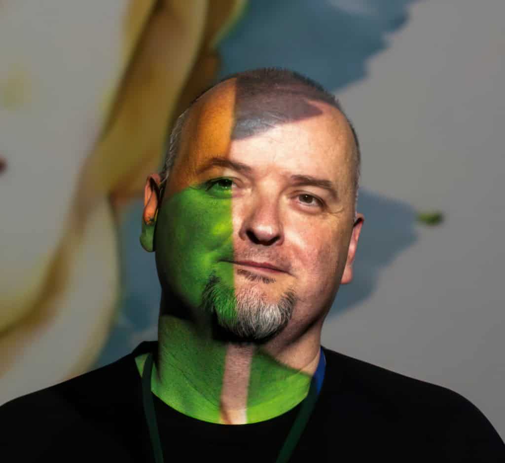 A man with a neutral expression stands in front of a colorful background. His face is partially lit with green and orange hues, creating a split-color effect. He has a goatee and wears a black shirt with a lanyard.