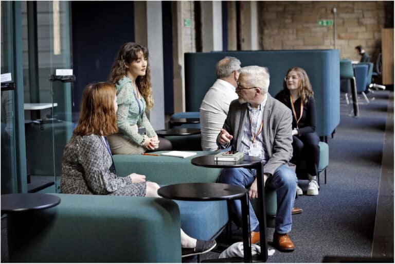 A group of people sitting in a modern lounge area, engaged in conversation. Some are seated on green sofas, while others are on chairs. The atmosphere is casual. Tables and glass walls are visible in the background.