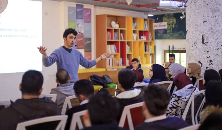 A man in a blue sweater is giving a presentation to a group of seated people in a classroom. The audience includes men and women, some wearing headscarves. Bookshelves and a chalkboard are in the background.