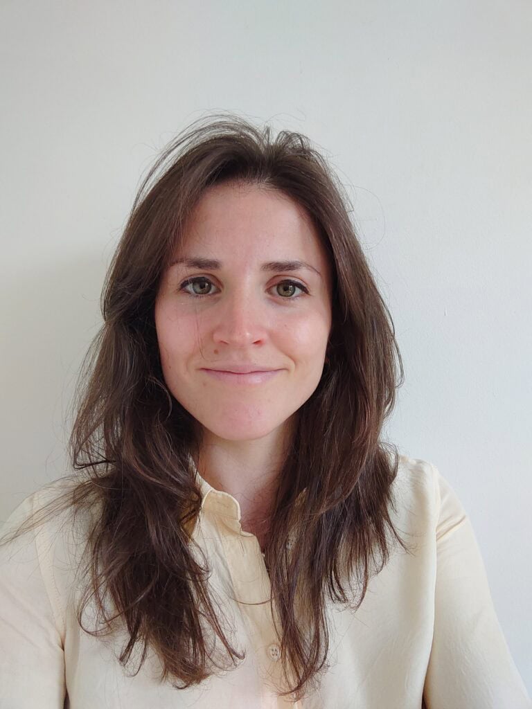 A person with long brown hair and wearing a cream-colored shirt smiles at the camera against a plain white background.