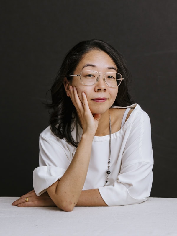 A person with long dark hair and glasses is leaning on a table with one hand on their cheek. They are wearing a white blouse and looking at the camera against a dark background.